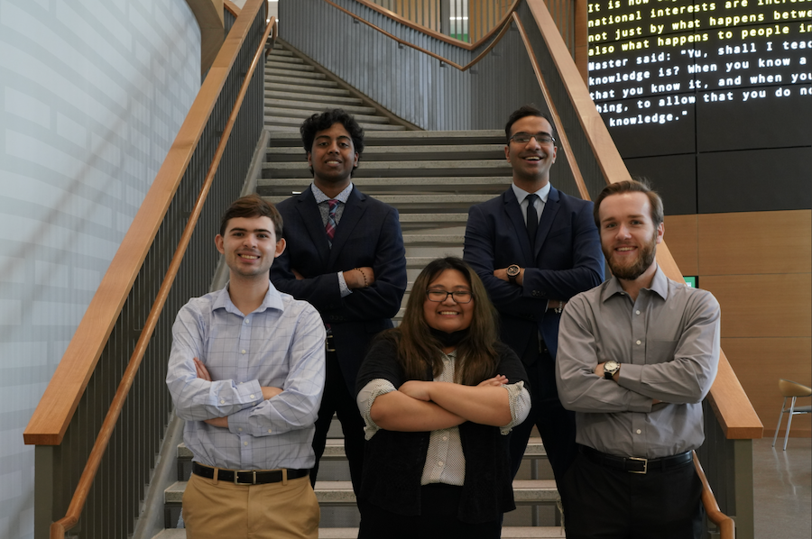 Five George Mason students who are members of HackOverFlow's Presidential Committee stand with their arms folded on a staircase in Horizon Hall. 