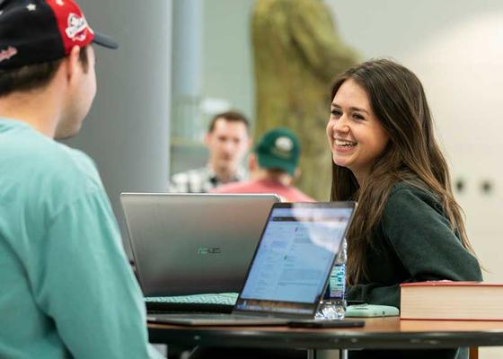 Female student smiles and converses with male student across a table filled with laptops and textbooks in the atrium at the Antonin Scalia School of Law at George Mason University